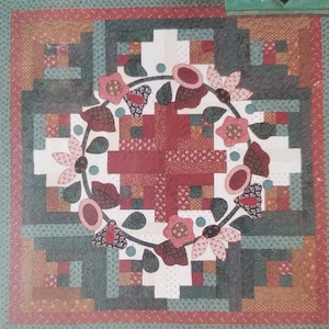 May include: A handmade quilt with a floral wreath design. The quilt features a central cross pattern in shades of red and white, surrounded by a wreath of flowers and leaves. The border is a mix of green, brown, and red fabrics, creating a cozy, handcrafted feel.