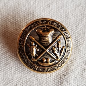 May include: A round, antique gold-colored button with a detailed embossed design. The central image features a crown, crossed oars, a turtle, and a canoe. The button has a textured border and is set against a neutral fabric background.