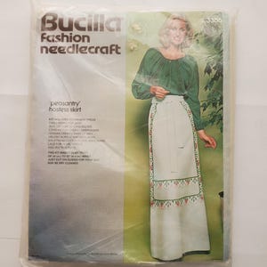 May include: A Bucilla Fashion Needlecraft kit for a 'peasantry' hostess skirt. The package shows a woman wearing the skirt, which has a white base with a floral embroidered border. The kit includes fabric, embroidery yarn, and instructions.