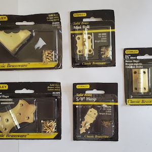 May include: An assortment of Stanley Classic Brassware hardware, including corner braces, mini bolts, narrow hinges, ornamental hinges, and a hasp. Each item is packaged in a black and yellow blister pack with the Stanley logo and product details.