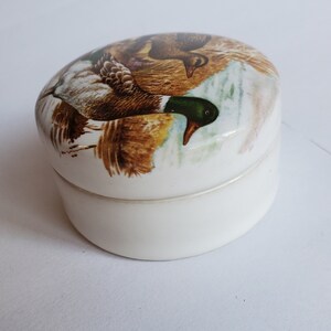 May include: A small, oval-shaped trinket box with a white base and lid. The lid features a detailed illustration of two ducks in a natural setting, with brown, green, and white feathers. The box is likely made of ceramic or porcelain.