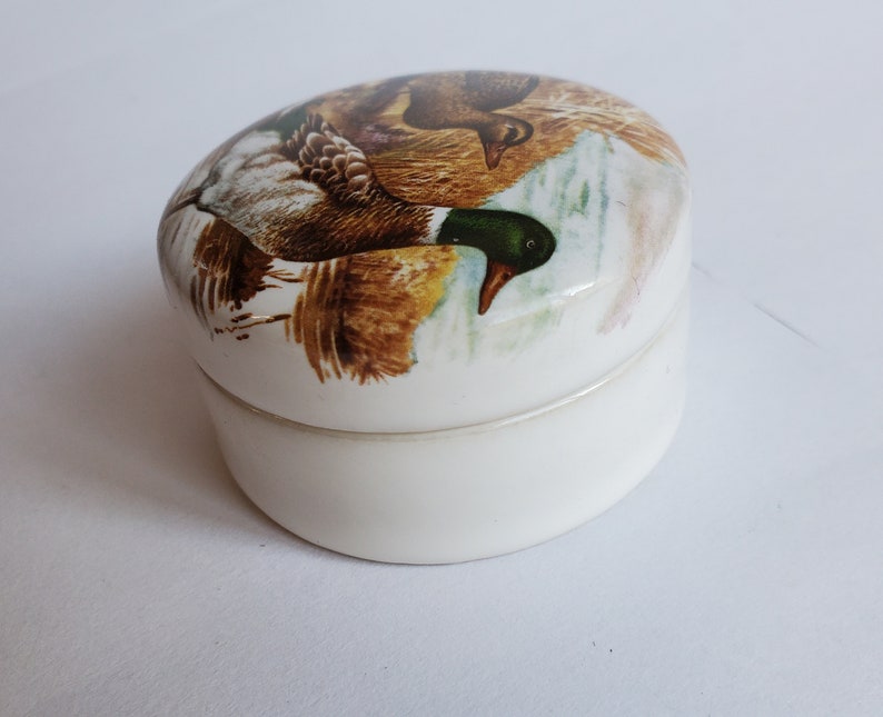 May include: A small, oval-shaped trinket box with a white base and lid. The lid features a detailed illustration of two ducks in a natural setting, with brown, green, and white feathers. The box is likely made of ceramic or porcelain.