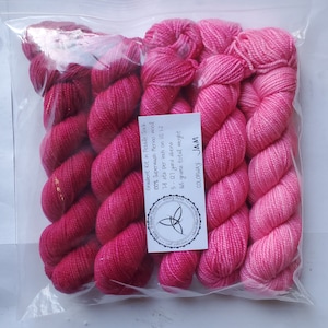 May include: A clear plastic bag containing a gradient yarn kit. The kit includes several skeins of pink yarn, ranging in shades from deep magenta to light pink. A white tag displays product details, including the yarn's composition and weight.