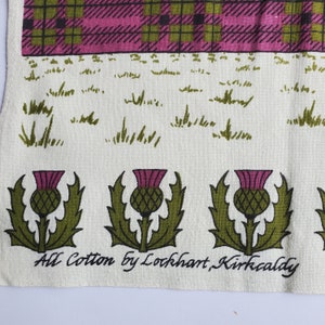 May include: Cream-colored textile with a tartan pattern in pink, green, and black at the top. Below is a field of green grass illustrations, and a row of green and purple thistle illustrations. The text "All Cotton by Lockhart, Kirkcaldy" is printed at the bottom.