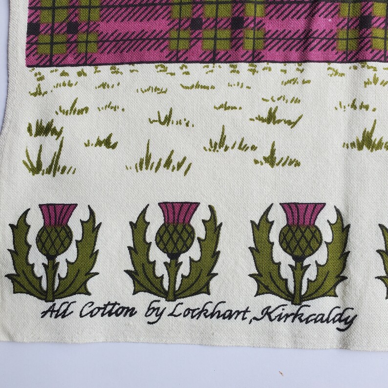 May include: Cream-colored textile with a tartan pattern in pink, green, and black at the top. Below is a field of green grass illustrations, and a row of green and purple thistle illustrations. The text "All Cotton by Lockhart, Kirkcaldy" is printed at the bottom.