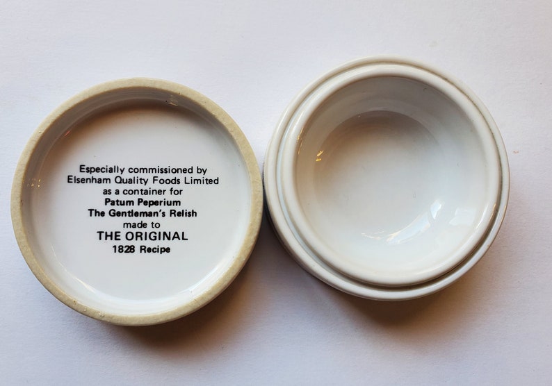 May include: Open, white ceramic container with a lid. The lid's interior features text: "Especially commissioned by Elsenham Quality Foods Limited...The Gentleman's Relish made to THE ORIGINAL 1828 Recipe."