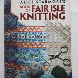 May include: A book titled "Alice Starmore's Book of Fair Isle Knitting" with a colorful knitted sweater design on the cover. Knitting needles, scissors, and balls of yarn in various shades of blue, green, and gray are also visible.