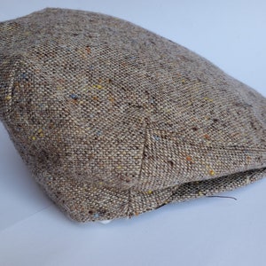 May include: A brown tweed flat cap with a classic design. The cap is made of a textured fabric with flecks of color, including yellow, blue, and orange. The hat is a traditional style, suitable for various occasions.