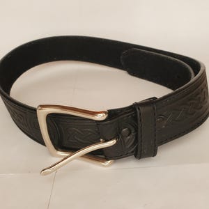 May include: A black leather belt with a silver-colored buckle. The belt has a Celtic knot pattern embossed along its length. The buckle is a square shape with a single prong. The belt is likely 1.5 inches wide.