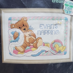 May include: A rectangular framed cross-stitch embroidery featuring a brown teddy bear holding a smaller bear and a white blanket. The text reads "EVAN'S NAPPING" in blue thread. A yellow duck, a block with the letter A, and a ball are also depicted.