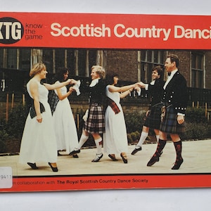 May include: A rectangular book cover with a bright red border titled KTG Know the Game Scottish Country Dancing. The cover features a photograph of six people in white dresses and kilts performing a dance in front of a stone building.