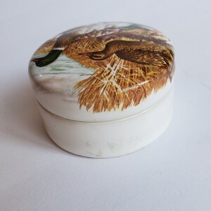 May include: Oval-shaped, white ceramic trinket box with a lid featuring a detailed illustration of two ducks in a natural setting. The ducks are brown, green, and white, set against a backdrop of reeds and water.