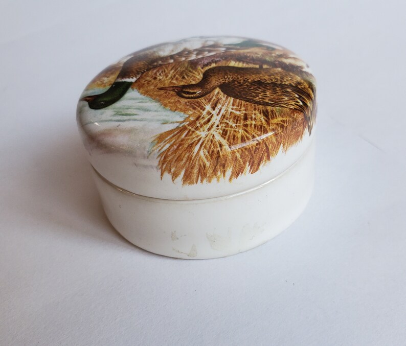 May include: Oval-shaped, white ceramic trinket box with a lid featuring a detailed illustration of two ducks in a natural setting. The ducks are brown, green, and white, set against a backdrop of reeds and water.