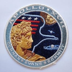 May include: Embroidered patch featuring the Apollo 17 mission. The patch has a blue border and depicts a gold bust, stars, and a stylized depiction of the moon and planets. The text "APOLLO 17" and astronaut names are visible.