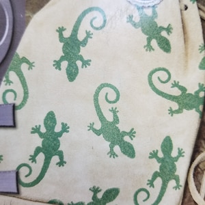 May include: A cream-colored suede pouch with a green lizard pattern. The drawstring bag features a fringe trim at the bottom and a decorative silver-colored button. The lizards are stylized with curled tails.