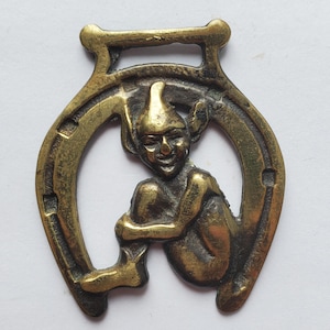 May include: A vintage brass horseshoe charm featuring a seated elf figure. The elf is depicted with pointed ears and a smiling face, nestled within the horseshoe's curve. The charm has a loop at the top for hanging.