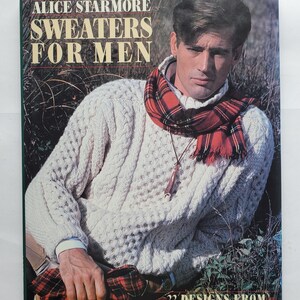 May include: A book cover featuring a man wearing a white cable-knit sweater and a red and black plaid scarf. The book title reads "SWEATERS FOR MEN" by Alice Starmore. The cover also includes the text "22 DESIGNS FROM THE SCOTTISH ISLES."