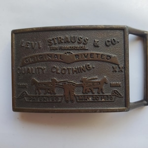 May include: A rectangular bronze-toned metal belt buckle featuring embossed text that reads Levi Strauss & Co. San Francisco, Cal. Original Riveted Quality Clothing XX. It includes a graphic of two horses pulling a pair of jeans and the date May 20 1873.