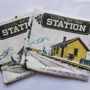 May include: Two off-white fabric tea towels featuring a printed illustration of a yellow train station. The text reads Down By The Station and Old South Canterbury, Connecticut. The design includes a train, tracks, and figures on a platform.