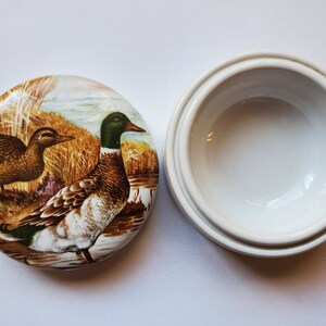 May include: A white ceramic trinket box with a lid featuring a detailed painting of two ducks in a natural setting. The ducks are brown, green, and white, with a backdrop of reeds and water.