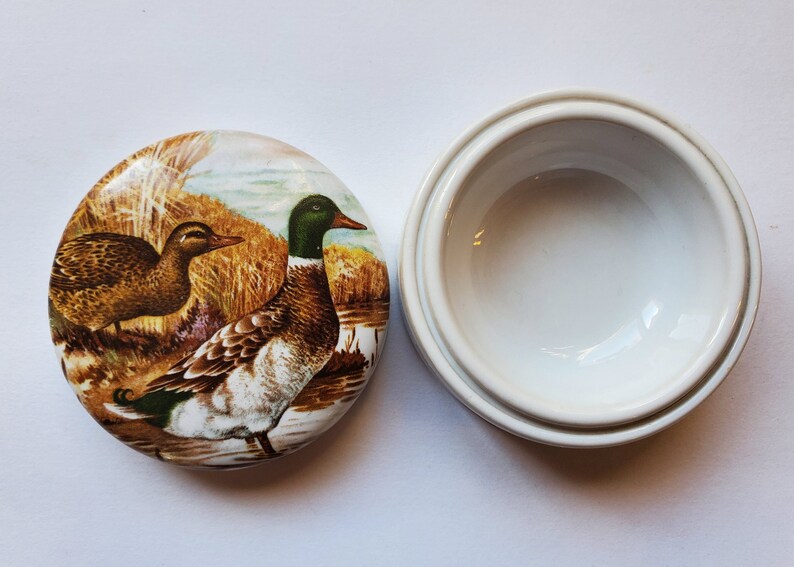 May include: A white ceramic trinket box with a lid featuring a detailed painting of two ducks in a natural setting. The ducks are brown, green, and white, with a backdrop of reeds and water.