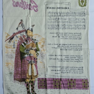 May include: A cream-colored textile featuring a Scottish theme. It displays a bagpiper in traditional attire, the word "Scotland", sheet music, and lyrics from "Amazing Grace". Thistle motifs border the top and bottom.