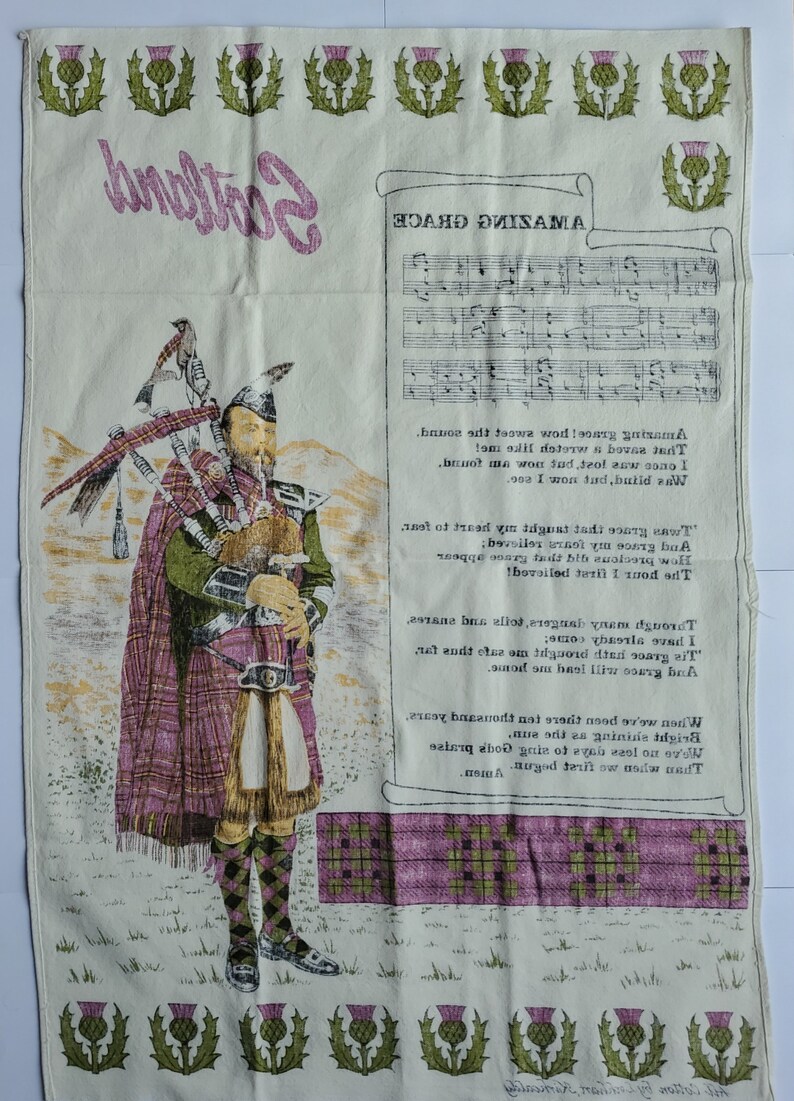 May include: A cream-colored textile featuring a Scottish theme. It displays a bagpiper in traditional attire, the word "Scotland", sheet music, and lyrics from "Amazing Grace". Thistle motifs border the top and bottom.