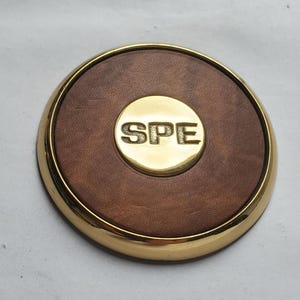 May include: A circular emblem featuring a polished gold-tone metal rim and a brown leather-like inlay. In the center, a smaller circular gold-tone metal disc is embossed with the letters SPE in a bold, sans-serif font, creating a layered design.