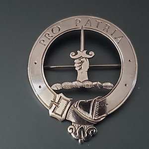 May include: A silver-toned brooch with the words "PRO PATRIA" encircling a central design. The design features a hand holding a sword, a shield, and a helmet. The brooch is likely a clan crest badge.