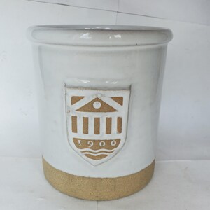 May include: A cylindrical ceramic crock with a glossy white glaze on the upper portion and an unglazed tan base. The front features a raised shield-shaped emblem in tan, displaying a building icon above the text 1900 and wavy lines below.