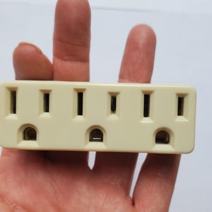 May include: A cream-colored electrical outlet adapter with three standard outlets. The adapter has a rectangular shape with four vertical slots and three pairs of horizontal slots. The adapter is held in a hand.