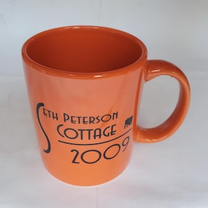 May include: A bright orange ceramic mug with a curved handle. The front features black text that reads Seth Peterson Cottage 2009, with a small black graphic of a building silhouette positioned to the right of the word Cottage.