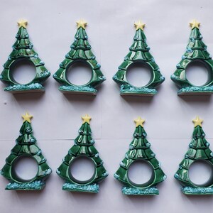May include: Eight ceramic Christmas tree napkin rings. Each tree is green with a yellow star on top and light blue accents. The napkin rings have a circular opening in the center.