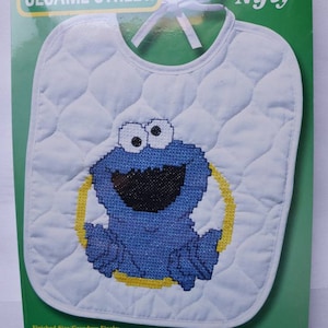 May include: A white quilted bib featuring a cross-stitch design of the Cookie Monster from Sesame Street. The bib measures 9.5 x 11 inches and has a yellow border around the character. The packaging includes the Sesame Street logo.