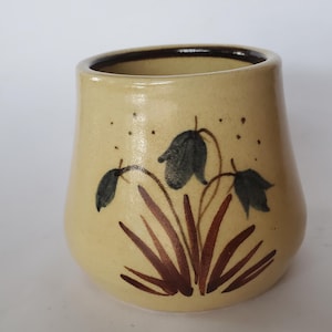 May include: A small, hand-painted ceramic vase with a light yellow base and a dark brown rim. The vase features a floral design with bluebell-like flowers and brown stems and leaves. The vase has a slightly rounded shape.