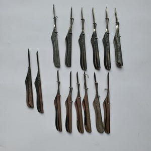 May include: An assortment of antique metal tools, possibly for leatherworking or similar crafts. The tools have a dark, aged appearance with a pointed blade and a curved handle. The collection includes various sizes and styles, arranged on a white surface.