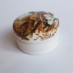 May include: A small, white ceramic trinket box with a lid featuring a detailed illustration of ducks in a natural habitat. The artwork showcases brown, green, and white feathers, set against a backdrop of reeds and foliage.