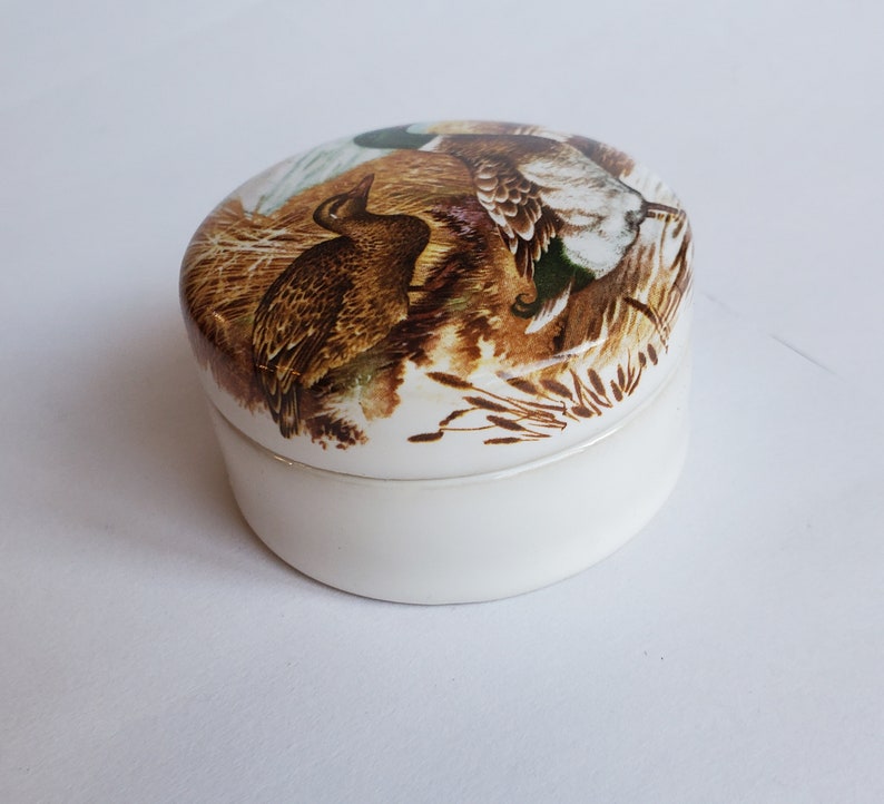 May include: A small, white ceramic trinket box with a lid featuring a detailed illustration of ducks in a natural habitat. The artwork showcases brown, green, and white feathers, set against a backdrop of reeds and foliage.
