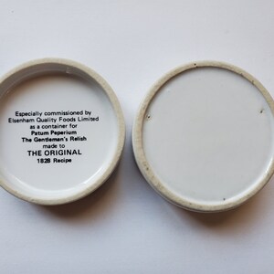 May include: Two small, round, white ceramic containers with a cream-colored rim. One container has text that reads "The Gentleman's Relish" and "1828 Recipe". The other container is empty.
