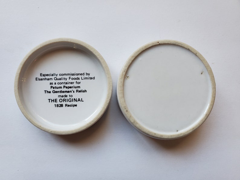 May include: Two small, round, white ceramic containers with a cream-colored rim. One container has text that reads "The Gentleman's Relish" and "1828 Recipe". The other container is empty.