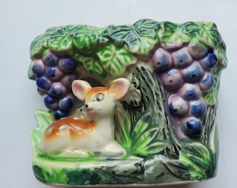 May include: A ceramic figurine featuring a fawn resting near a tree with grapes. The fawn has white fur with brown spots and a brown back. The grapes are purple and the leaves are green. The base is green.