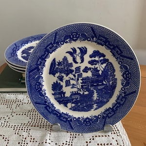 May include: A set of round ceramic plates featuring a classic blue and white willow pattern. The design depicts a detailed landscape with pagodas, a bridge, willow trees, and two birds in flight, framed by a decorative geometric border pattern.