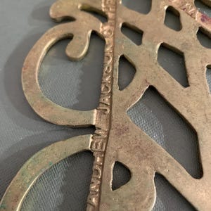 Op de afbeelding: Close-up van een vintage messing decoratief stuk met een sierlijk, opengewerkt ontwerp. De tekst "ANDREW JACKSON & THE" is zichtbaar, samen met "&copy; 1805". Het messing heeft een patina met hints van groen en blauw.