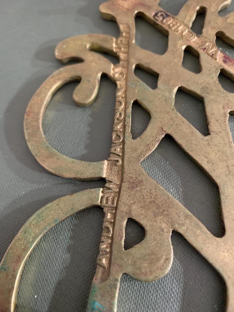 Op de afbeelding: Close-up van een vintage messing decoratief stuk met een sierlijk, opengewerkt ontwerp. De tekst "ANDREW JACKSON & THE" is zichtbaar, samen met "&copy; 1805". Het messing heeft een patina met hints van groen en blauw.