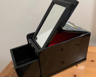 May include: A black, lacquered jewelry box with a hinged lid and a pull-out drawer. The lid is open, revealing a mirror and a red velvet interior. The box is on a wooden surface.