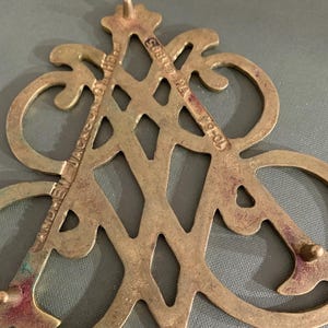 Op de afbeelding: Een vintage decoratief stuk van messing met een sierlijk ontwerp. Het stuk bevat de tekst "ANDREW JACKSON HOTEL" en "NY &copy;1905 70-54". Het messing heeft een aangetaste patina, wat bijdraagt aan de antieke charme.