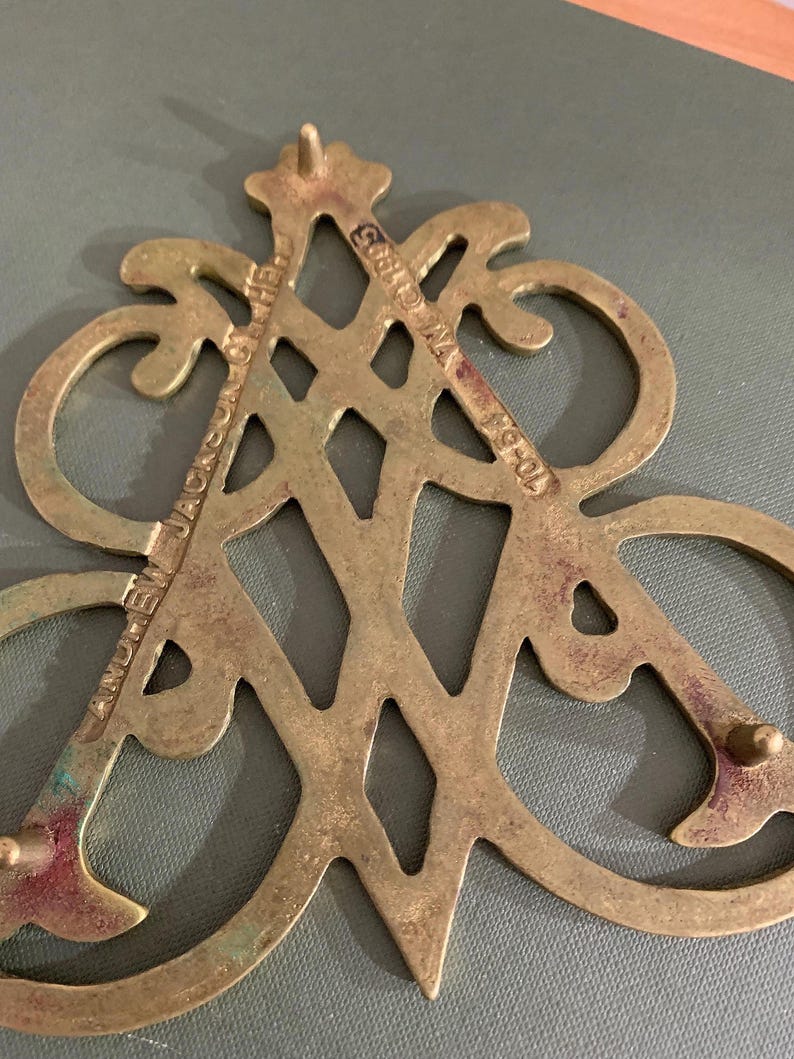 Op de afbeelding: Een vintage decoratief stuk van messing met een sierlijk ontwerp. Het stuk bevat de tekst "ANDREW JACKSON HOTEL" en "NY &copy;1905 70-54". Het messing heeft een aangetaste patina, wat bijdraagt aan de antieke charme.