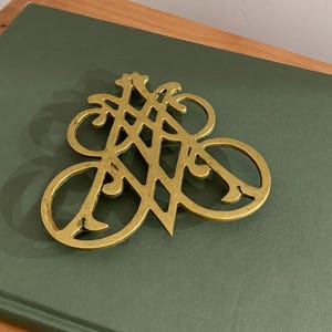 Op de afbeelding: Een decoratieve messing onderzetter met een open ontwerp. De onderzetter heeft een stervormig element en een reeks gebogen lijnen en lussen. Het ligt op een groene ondergrond.