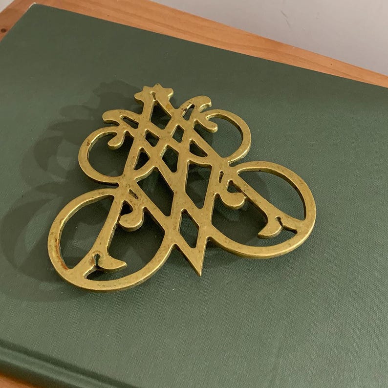 Op de afbeelding: Een decoratieve messing onderzetter met een open ontwerp. De onderzetter heeft een stervormig element en een reeks gebogen lijnen en lussen. Het ligt op een groene ondergrond.