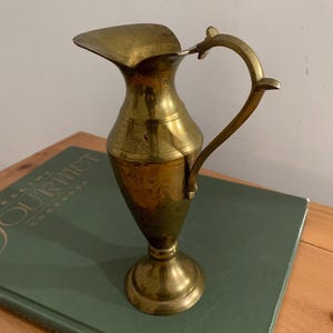 May include: A vintage brass pitcher with a curved handle and spout. The pitcher has a round base and a slender body, with a tarnished finish. The pitcher is approximately 7 inches tall.